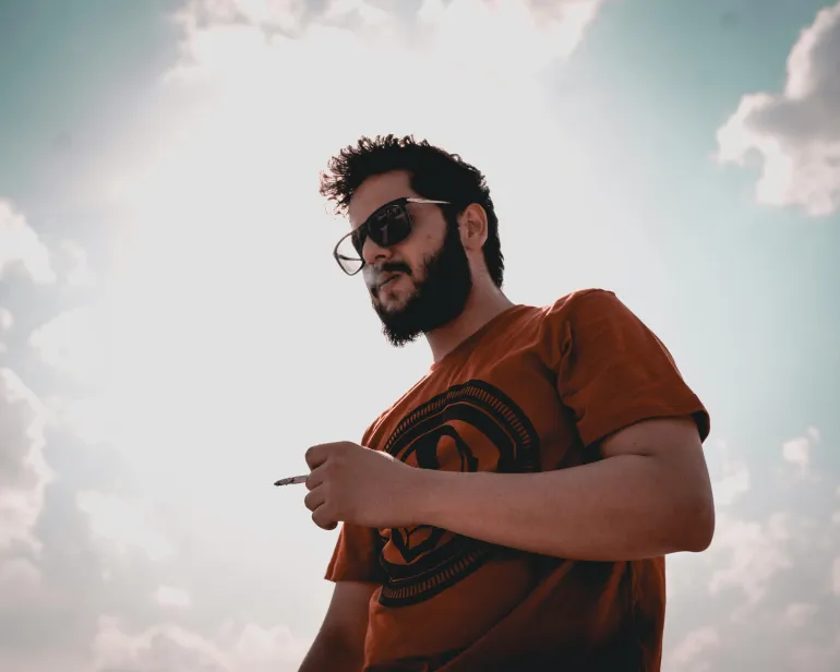 A bearded man wearing sunglasses smokes a cigarette under a bright sky.