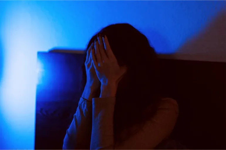 A moody and evocative image of a person covering their face in distress, captured in low light with deep blue and orange tones. The photograph conveys a sense of emotional tension, isolation, or internal turmoil. Ideal for illustrating concepts related to mental health, anxiety, depression, loneliness, or difficult emotions in visual storytelling.