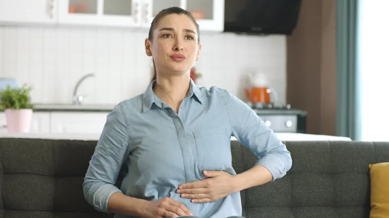 Young woman suffering from stomach spasm, abdominal pain at home. Stressed young woman massaging her belly on sofa, having stomach ache. Gastritis, abdominal or menstrual pain, diarrhea concept.