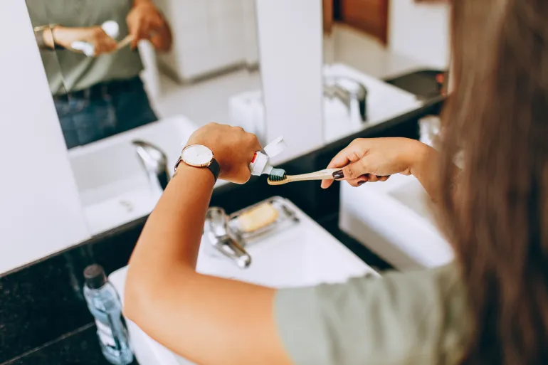 A woman is applying toothpaste to a toothbrush in a bathroom setting, showing daily oral hygiene habits.