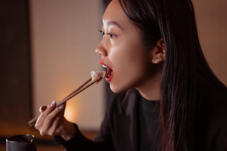 Asian woman savoring a dumpling using chopsticks, captured in a warm indoor setting.