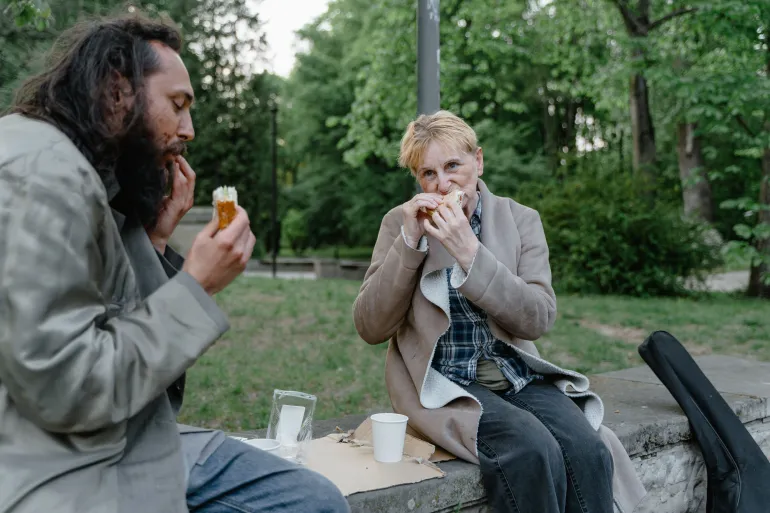 Two individuals share a meal outdoors on a park bench surrounded by greenery.