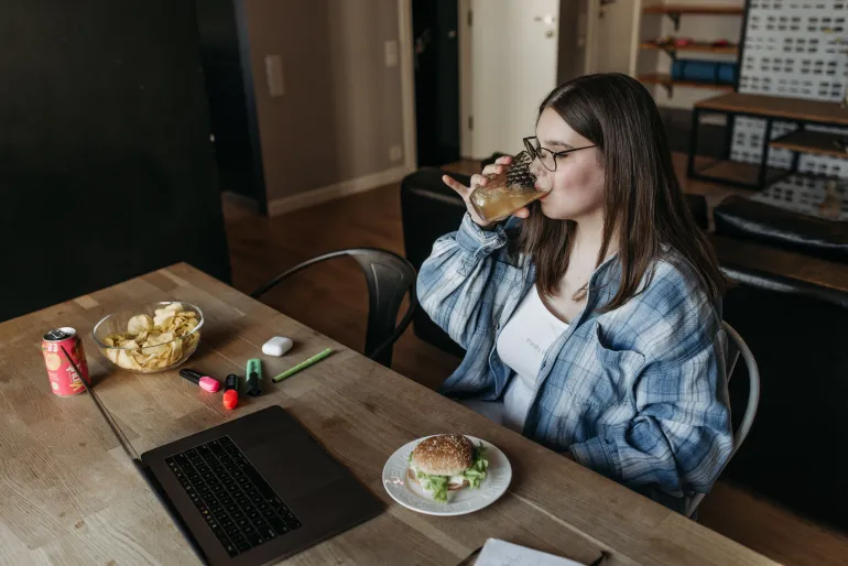 A young woman drinks from a glass while working on her laptop with snacks nearby.