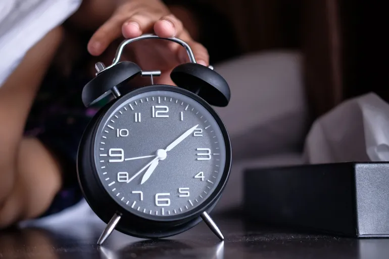 A hand reaches out to turn off an analog alarm clock in the early morning.