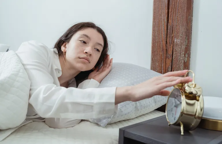 A woman in pajamas reaches to turn off an alarm clock in a cozy bedroom setting.
